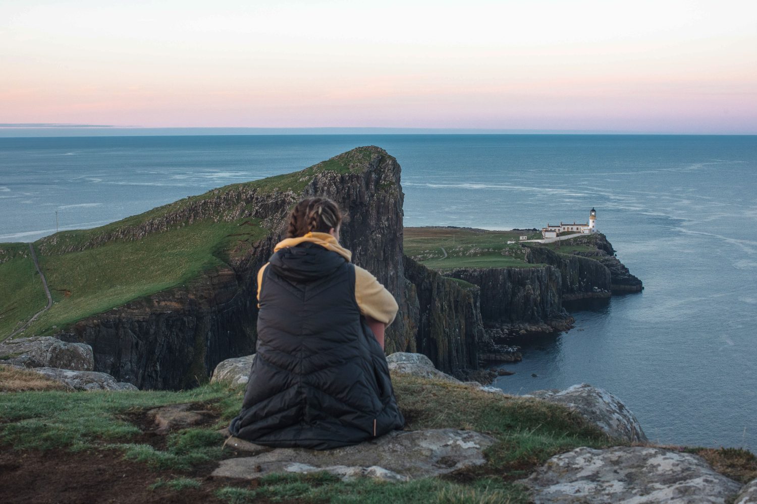 Travelers walking the scenic trail to Neist Point Lighthouse on the Isle of Skye, Scotland, with panoramic ocean views at sunset.